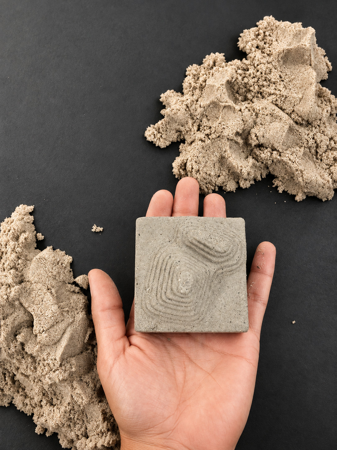 Mano sosteniendo una pieza de concreto con relieve topográfico, junto a arena cinética
