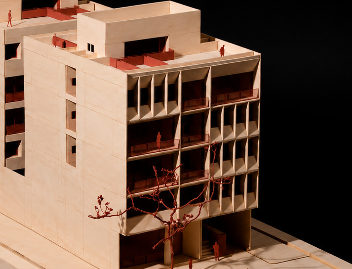 Maqueta de dos volúmenes de madera clara con terrazas en azotea, balcones rojos, un árbol rojo junto al acceso y figuras humanas, sobre fondo oscuro.