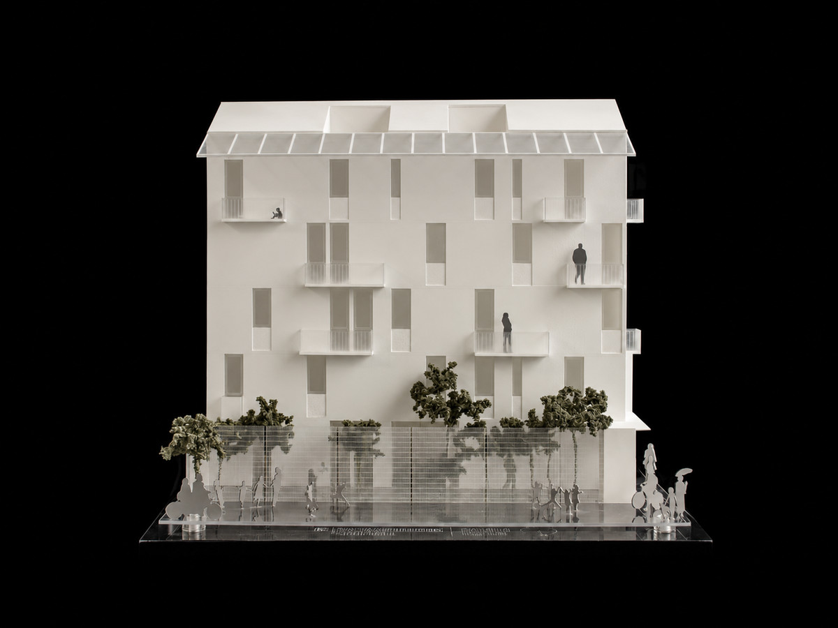 Maqueta frontal de un edificio blanco de cuatro niveles con balcones, vanos verticales, árboles y figuras a escala sobre una base de acrílico, fotografiada sobre fondo oscuro.